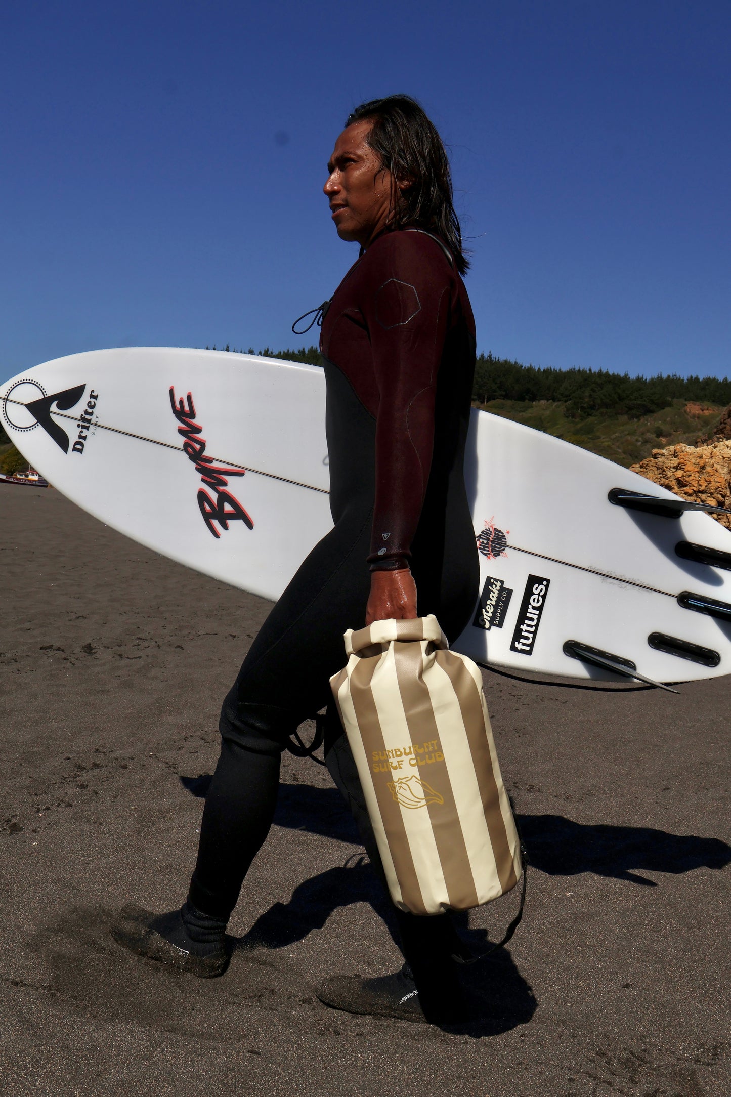 surfer wearing a wetsuit holding a printed dry bag and surf board